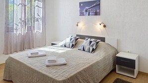 A neatly made bed with a patterned bedspread, two pillows, and two folded towels. There is a nightstand with a drawer, a wall-mounted light, and a framed picture above the bed.