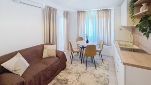 A compact living space with a brown sofa, a dining table with chairs, a small kitchen area, and a window with curtains. - Apartment Grozdana No.3 - with Balcony (Baska)