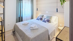 Ein ordentlich bezogenes Bett mit Kissen und gefalteten Handtüchern, ein Nachttisch und eine hängende Pflanze.