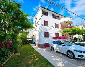 Un edificio blanco de varios pisos con balcones, un camino de entrada pavimentado y un coche blanco estacionado al frente.