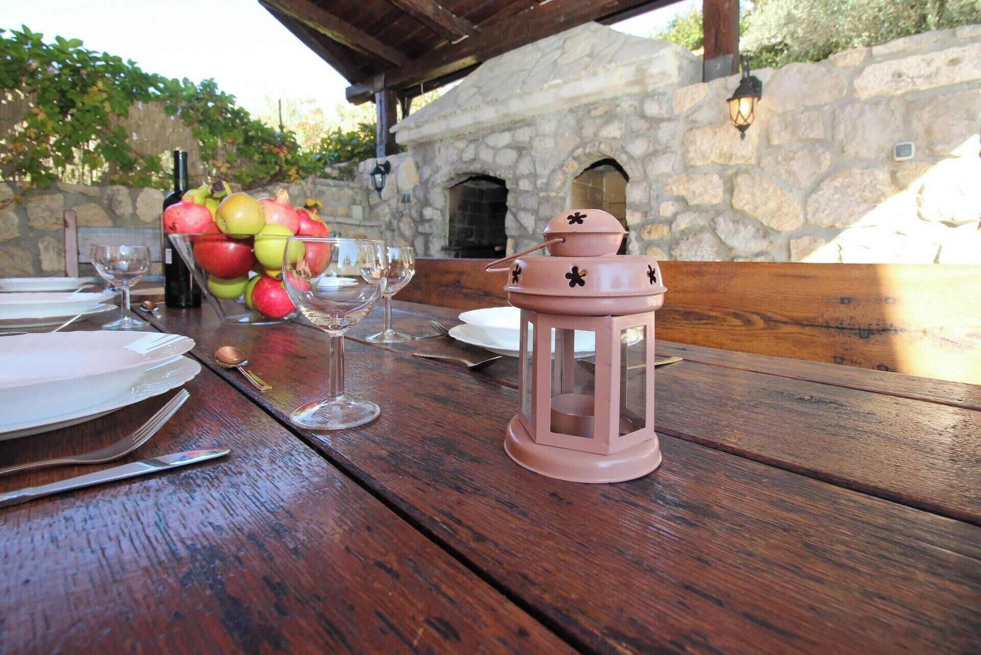A wooden table set for a meal with plates, cutlery, and wine glasses. A pink lantern and a bowl of fruit are also visible.