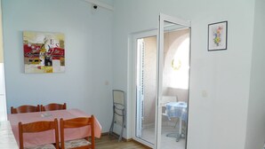 A dining area with a wooden table and chairs, a sliding glass door leading to a balcony, and a small table with a blue and white checkered tablecloth.