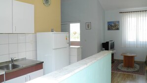 A compact kitchen with white cabinets, a built-in sink, and a refrigerator. There's a small television and a window with curtains.