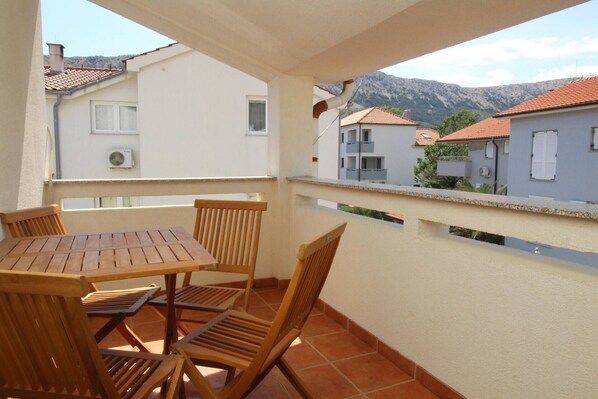 A balcony with a wooden table and chairs, overlooking a residential area with terracotta roofs and a mountain in the background.