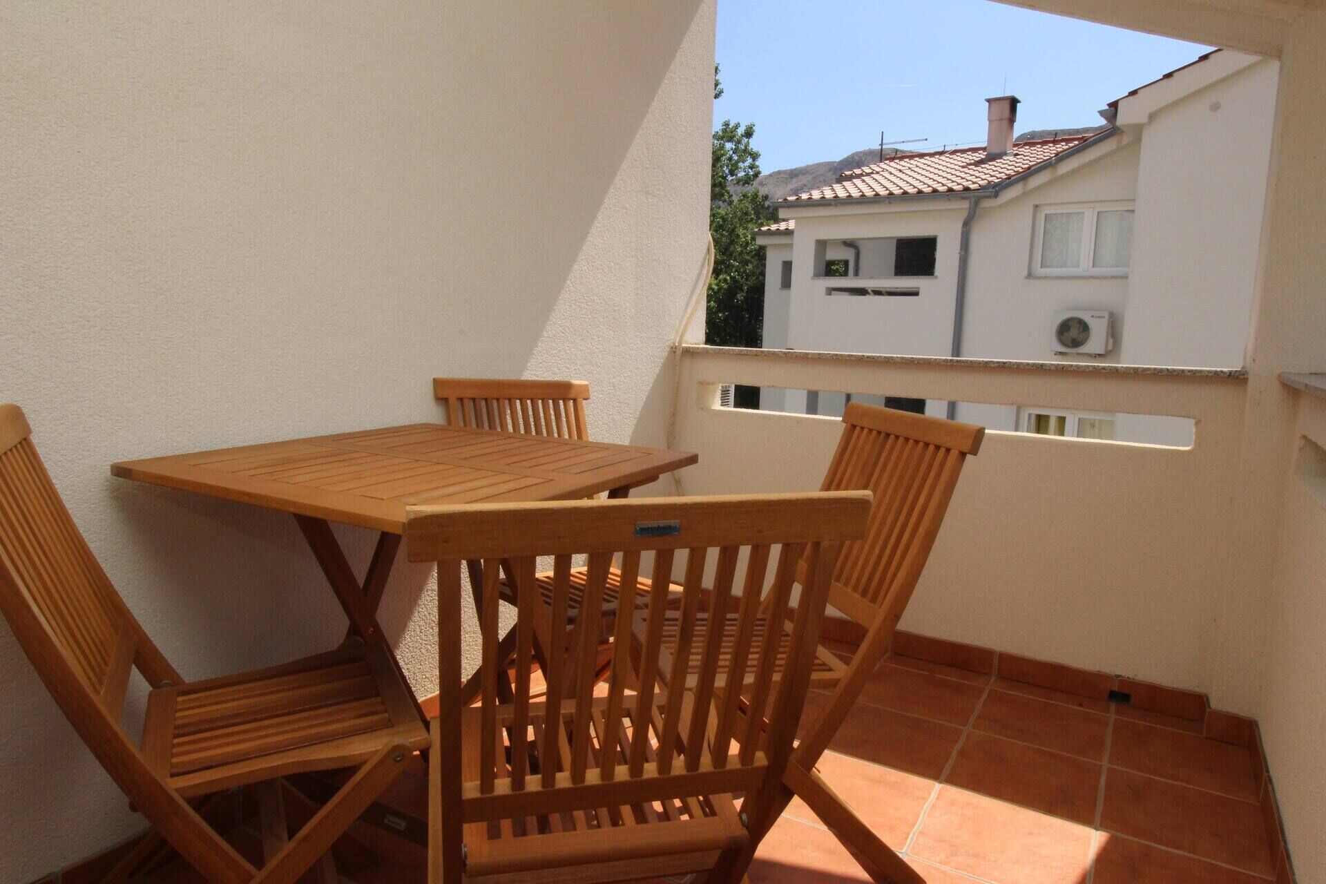 A balcony with a wooden table and chairs, a white wall, and a view of a building with a tiled roof.