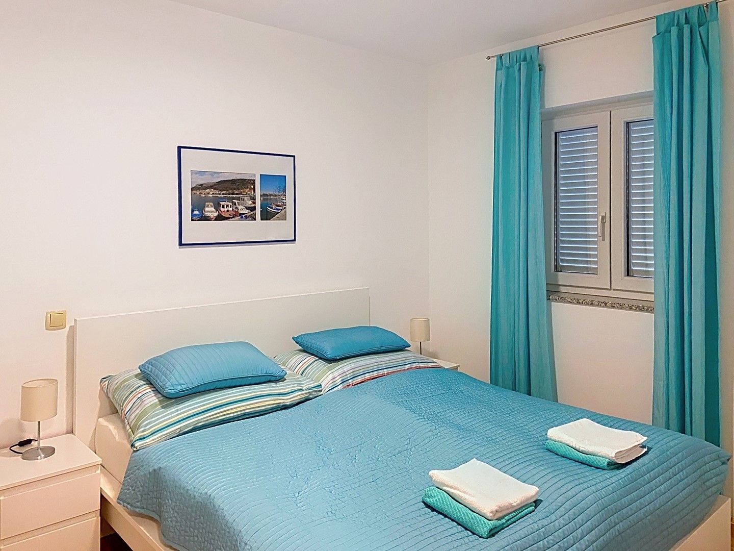 A neatly made bed with blue bedding and pillows, a bedside lamp, and a framed picture on the wall.