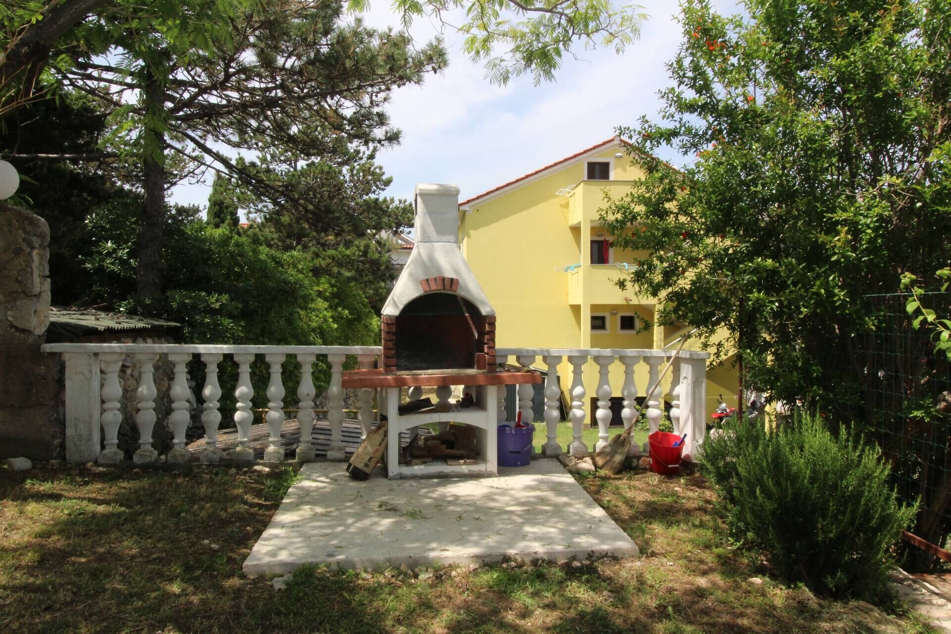 An outdoor brick oven in a garden with a white fence, a yellow building in the background, and greenery around.