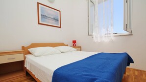 A single bed with a wooden headboard, a white mattress, and a blue blanket. There is a nightstand with a red lamp, a wooden drawer, and a framed picture on the wall. - Apartment Lovincic No.7 - with Sea View, 200 m from the Beach (Baska)