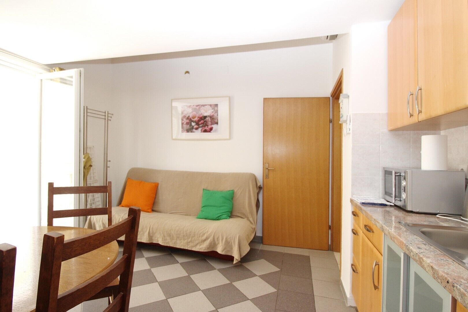 Een ruimtelijk ingerichte leefruimte met een kitchenette, een bank met kussens, een eettafel met stoelen, en een deur naar een andere kamer.