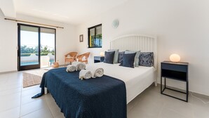 A modern bedroom with a large bed, a bedside table with a lamp, a view of the outdoors through a sliding door, and a small seating area with a chair and a table. - Casa Federica | 4 Bedrooms | Magnific Views | Loulé (Santa Bárbara de Nexe)