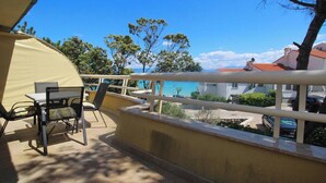 A balcony with a table and chairs, overlooking a coastal area with buildings and trees. - Apartment Mrkus brdo - with Balcony, near the Beach (Baska)