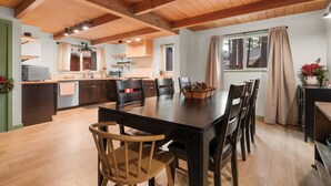 A spacious dining area with a wooden table and chairs, a kitchen in the background, and a window with curtains. - Charming Cabin Escape (Big Bear Lake)