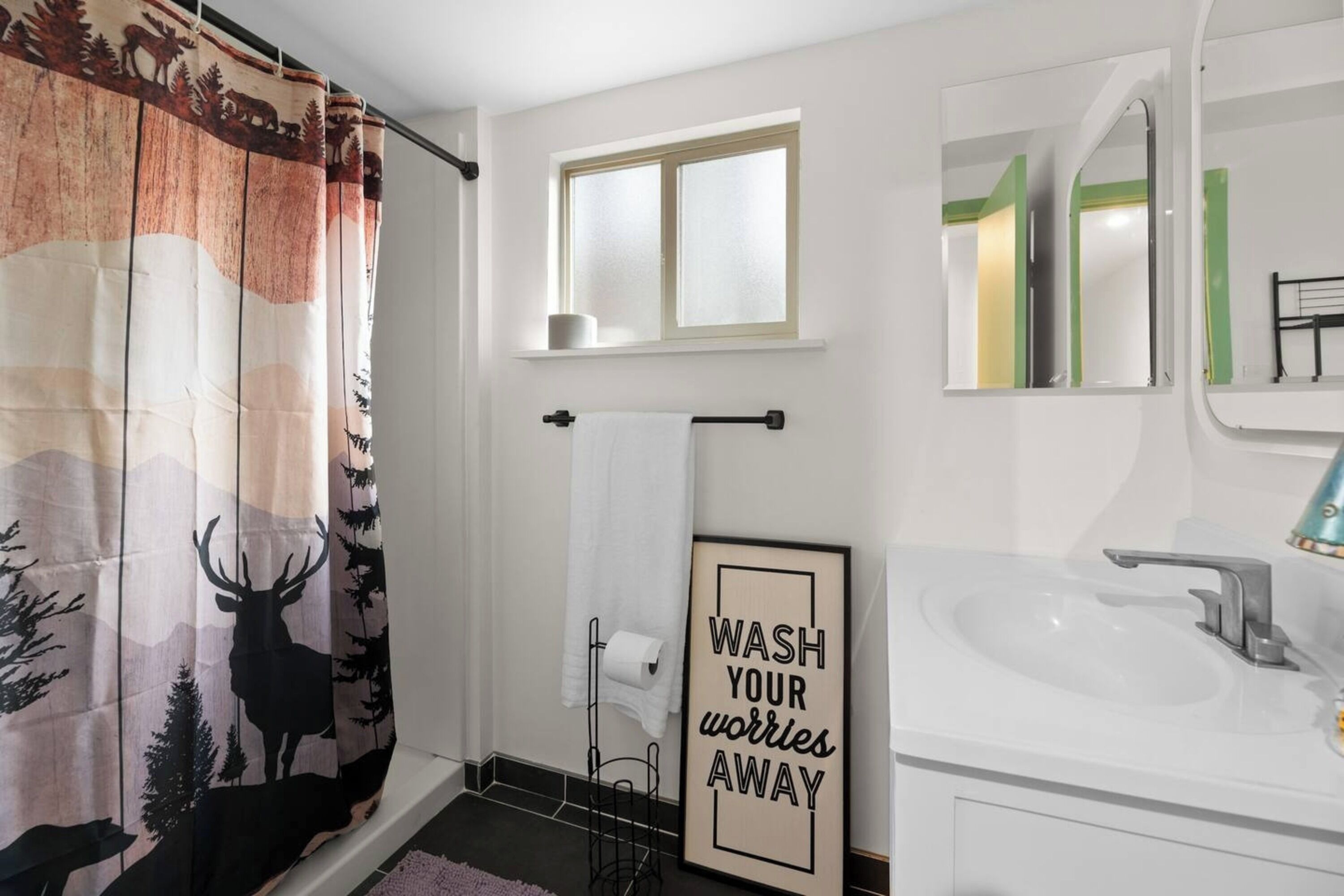 A bathroom with a shower curtain featuring a forest scene, a framed motivational quote on the wall, and a window above the sink.