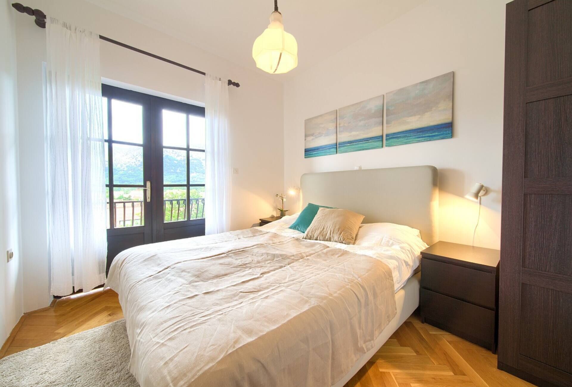 A bedroom with a bed, bedside tables, a nightstand, a lamp, a painting, and a view through a window.