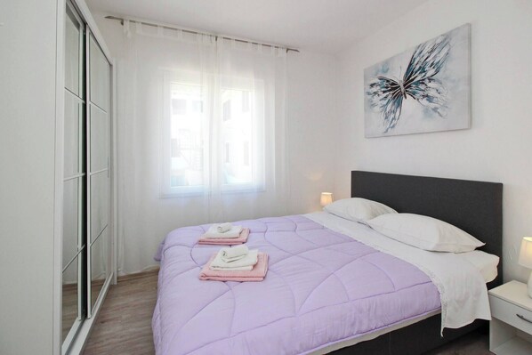 A bedroom with a bed, a nightstand, a wall-mounted artwork, and a window with sheer curtains.