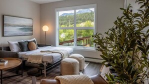 A bedroom with a bed, a bedside table, a lamp, a window with a view of greenery and mountains, and a painting on the wall. - Cosmo’s Getaway | Mtn Views | AC | BBQ | King Bed (Golden)
