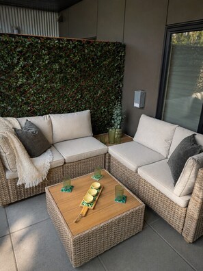 A modern outdoor seating area with a wicker sofa, a wooden coffee table with bowls and glasses, and a green leafy wall.