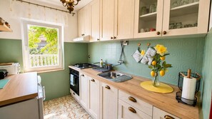 A kitchen with a gas stove, sink, wooden cabinets, and a window with a view of greenery. - Small semi-detached house Radojevic for 4-5 persons (Baska)
