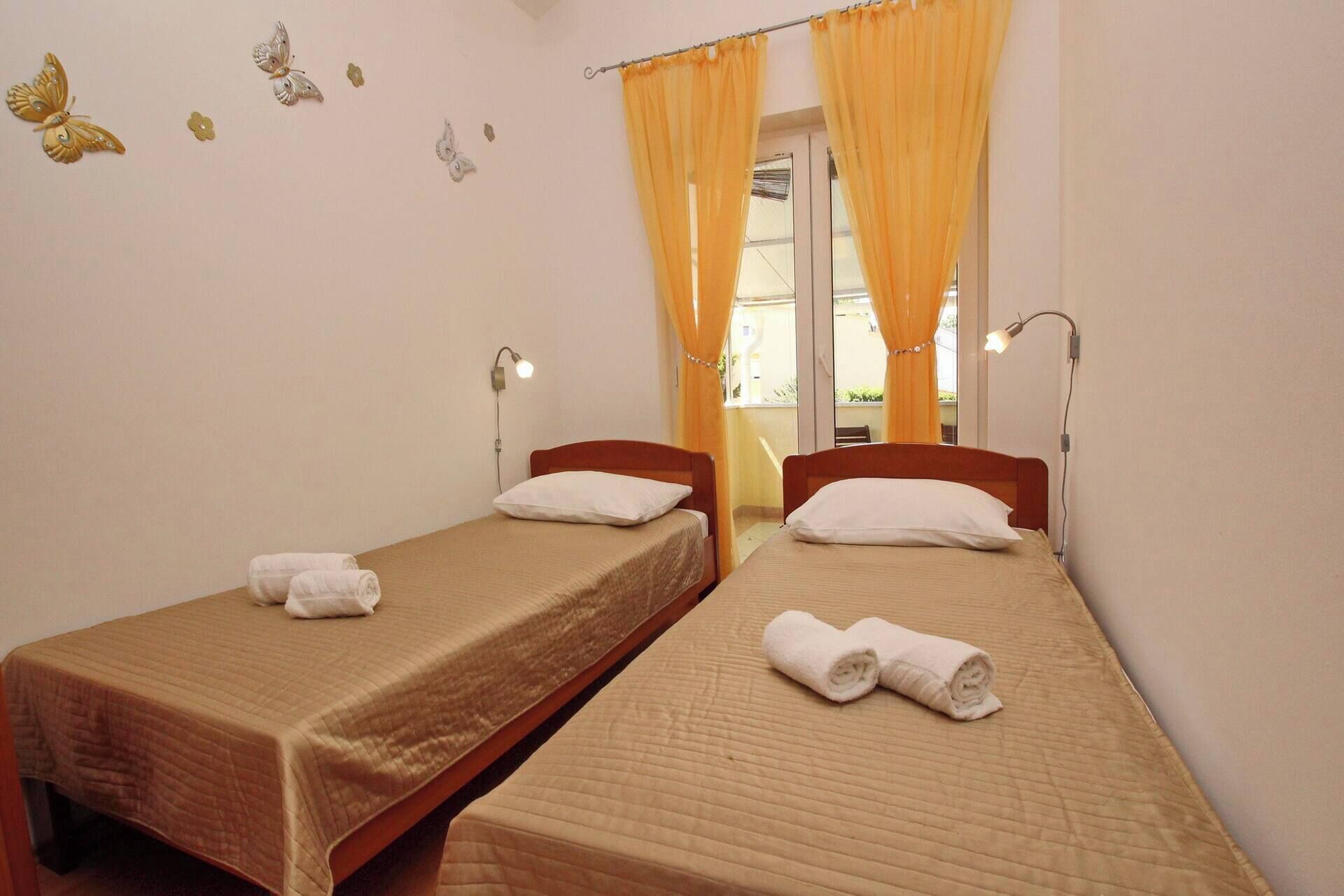 A room with two single beds, each with a rolled towel, a wooden headboard, and a white pillow. There are two wall-mounted lamps, a window with yellow curtains, and a view of an outdoor balcony.