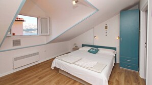 Une chambre avec un plafond en pente, un lit avec un matelas blanc et un oreiller turquoise, une table de chevet avec une lampe, et une vue sur une montagne enneigée par la fenêtre.