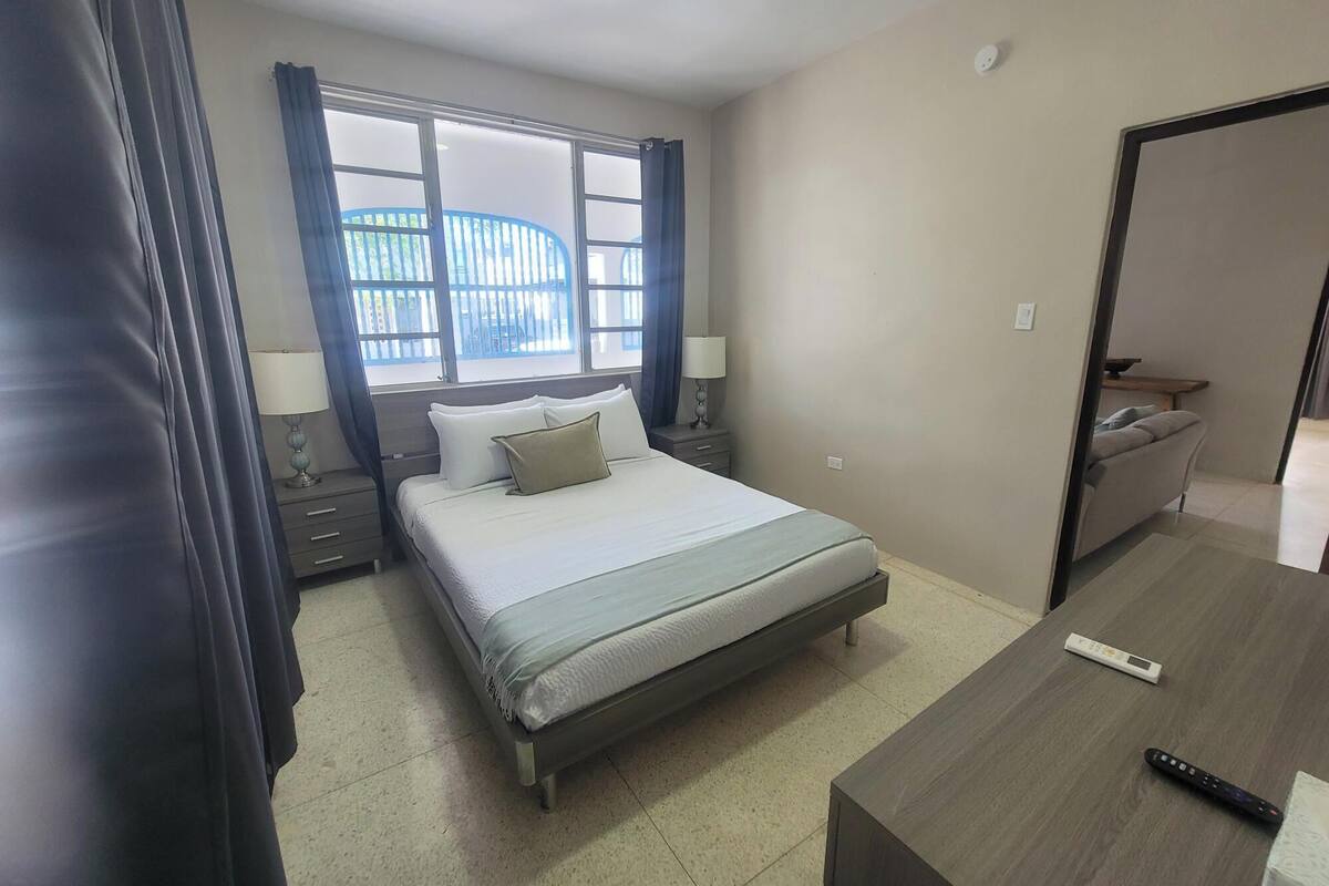 Habitación de hotel con cama, mesitas de noche, escritorio con control remoto y ventana con cortinas.