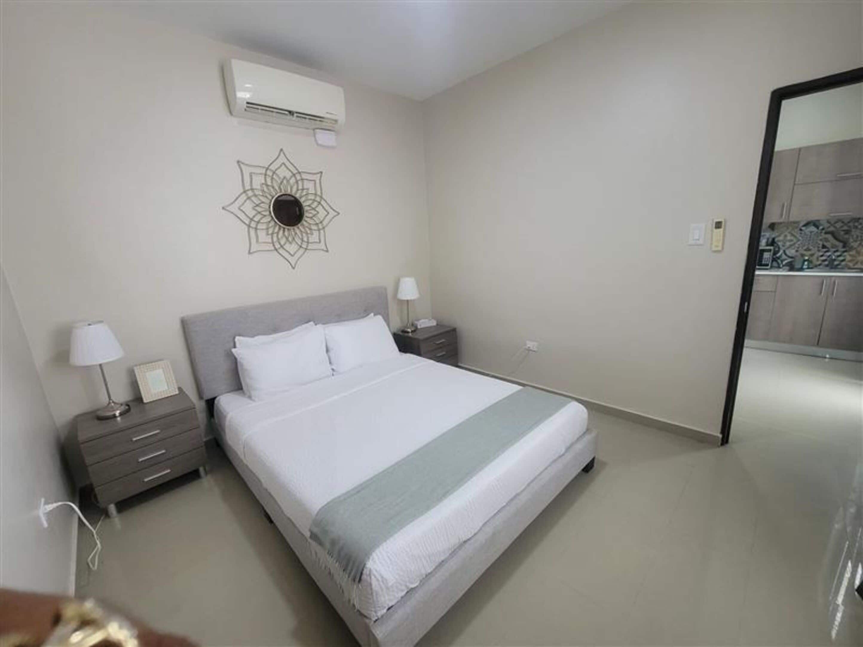 A bedroom with a bed, bedside tables, a wall-mounted air conditioner, and a decorative wall piece.