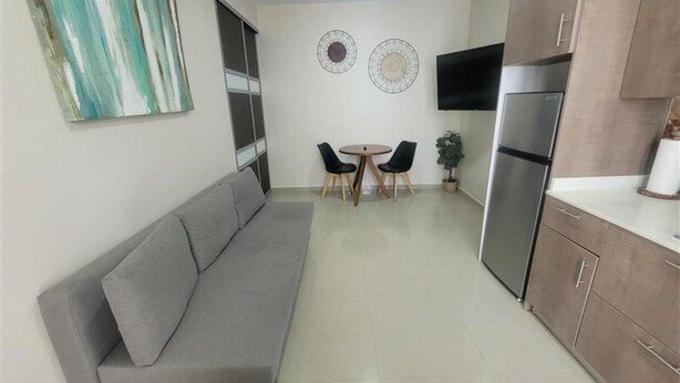 Cocina compacta con un sofá gris, una mesa de comedor con sillas, un refrigerador y un televisor montado en la pared.