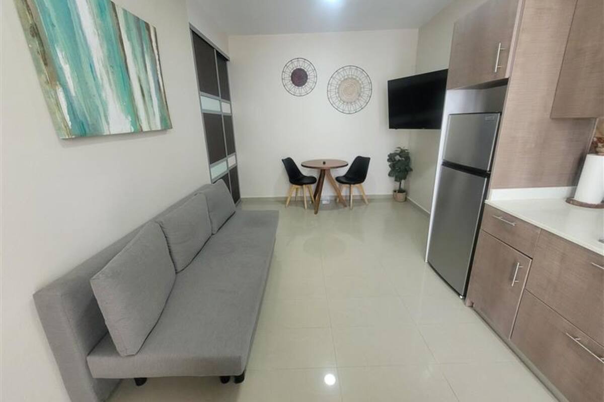 Cocina compacta con un sofá gris, una mesa de comedor con sillas, un refrigerador y un televisor montado en la pared.