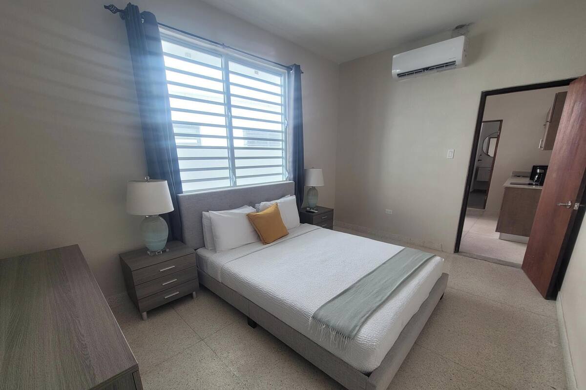Un dormitorio con cama, mesitas de noche, lámpara, ventana con cortinas, equipo de aire acondicionado y puerta que da a un baño.