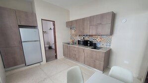 Cocina compacta con refrigerador, microondas y cafetera. Hay una mesa de comedor con sillas y una pequeña planta en maceta.