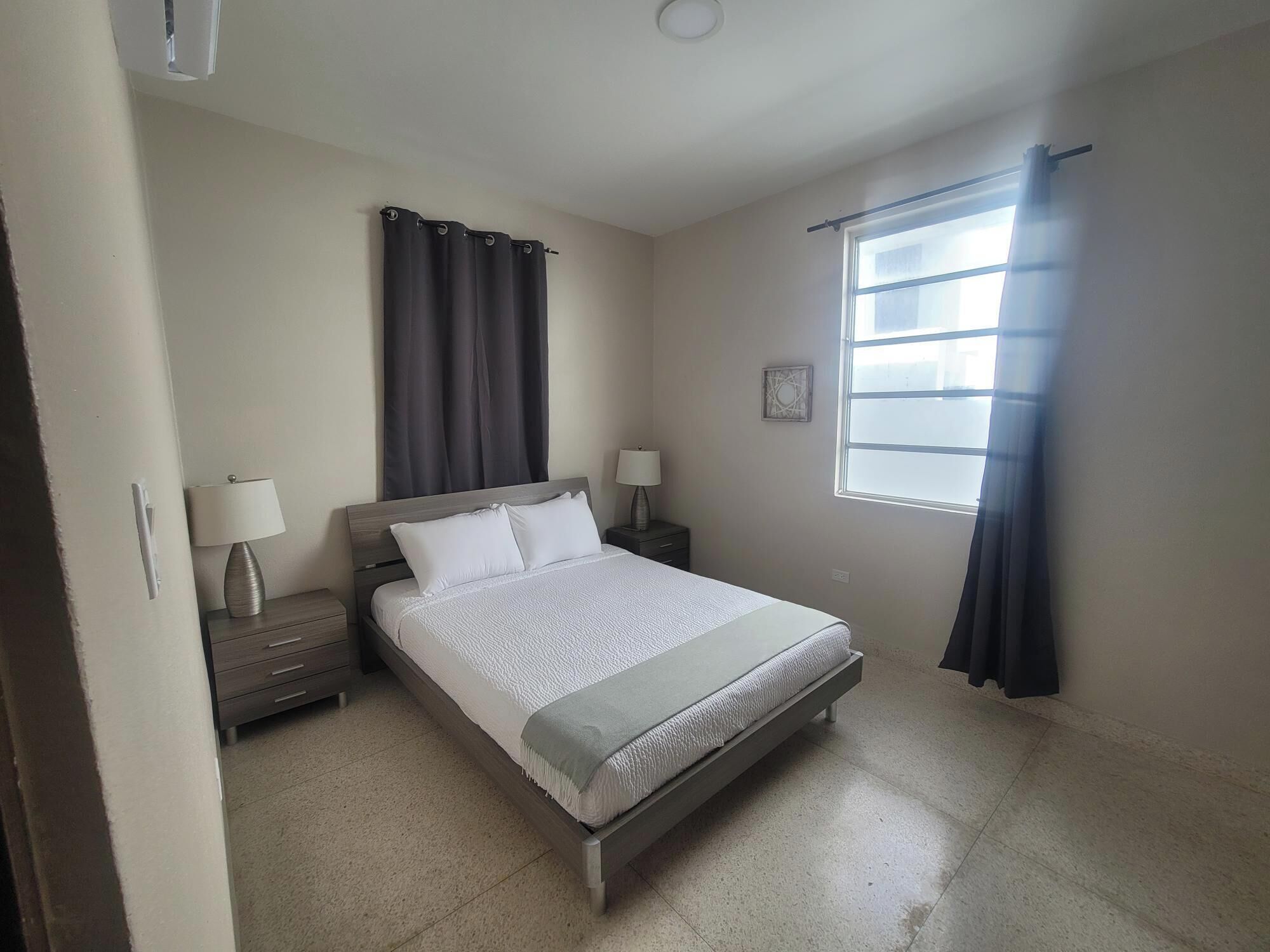 Un dormitorio con una cama, dos mesitas de noche con lámparas, una ventana con cortinas y un pequeño adorno en la pared.