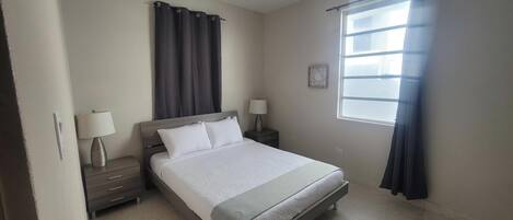 Un dormitorio con una cama, dos mesitas de noche con lámparas, una ventana con cortinas y un pequeño adorno en la pared.