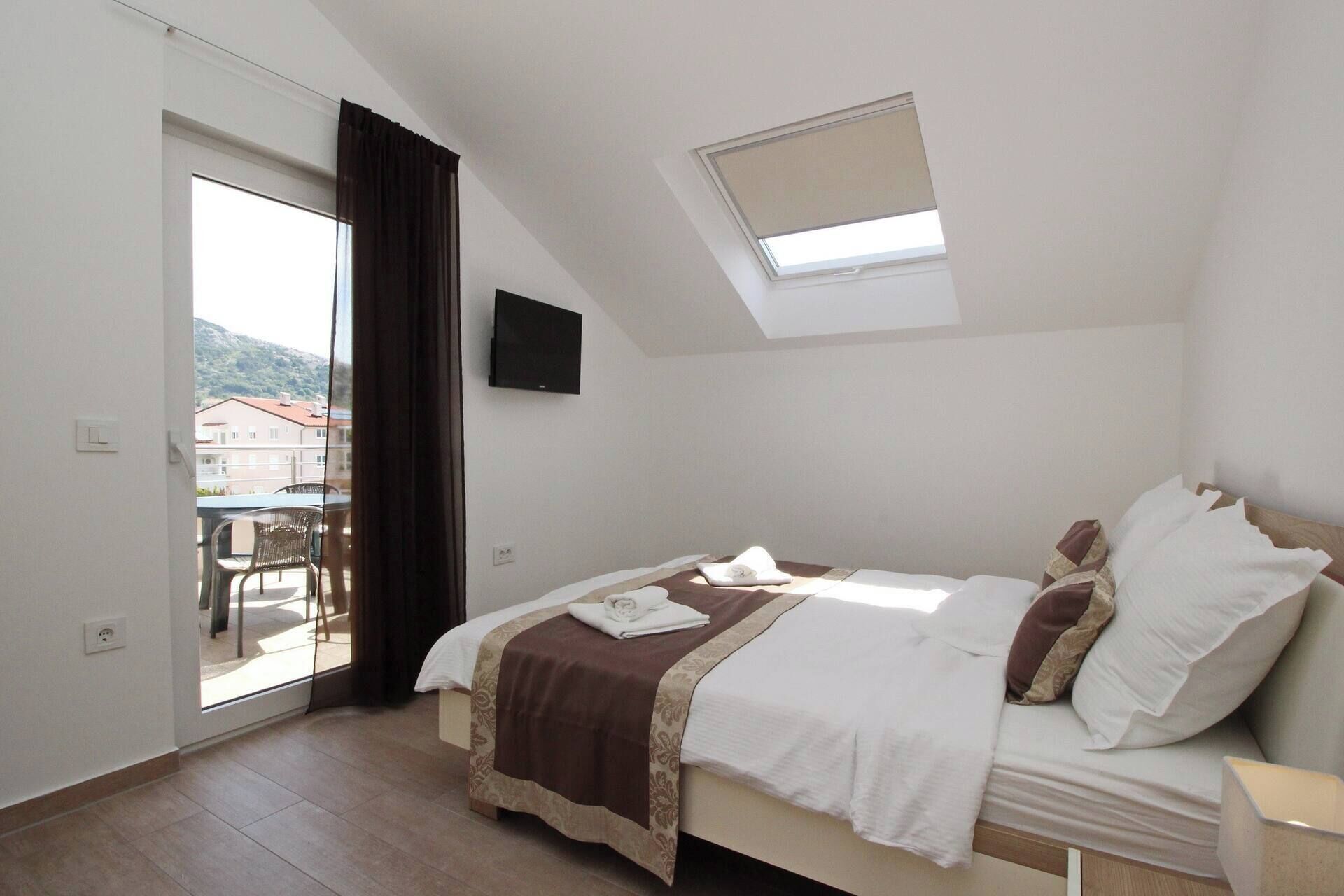 A bedroom with a bed, a TV, a balcony, and a view of buildings outside.