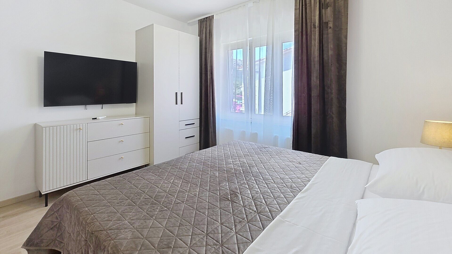 A hotel room with a bed, a TV mounted on the wall, a wardrobe, and a window with curtains.