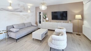Un salon moderne avec un canapé gris, une table basse blanche et un fauteuil blanc. Un téléviseur à écran plat est fixé au mur, et une lampe de sol ainsi qu’un vase décoratif sont posés sur la commode.