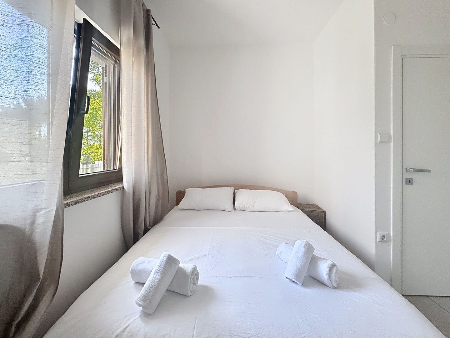 Ein Hotelzimmer mit einem Bett, zwei Kissen, einem Nachttisch, einem Fenster mit Vorhängen und einer Tür.