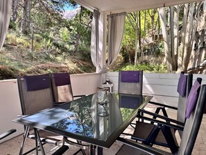 An outdoor patio with a glass table and chairs, surrounded by curtains and a view of trees and mountains.