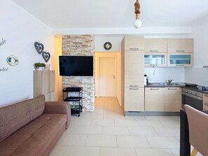 A modern living room with a sofa, a flat-screen TV mounted on a stone wall, and a kitchen with light wood cabinets and a stove.