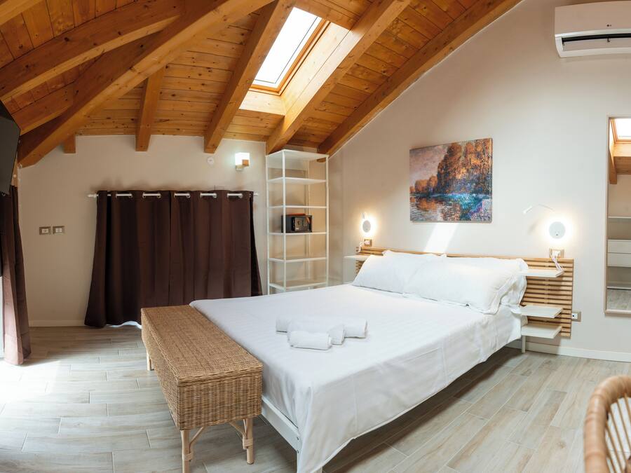Un dormitorio con techo de madera, un tragaluz, una cama con sábanas blancas, una mesita de noche con lámpara, un estante con un cuadro y un banco de mimbre.