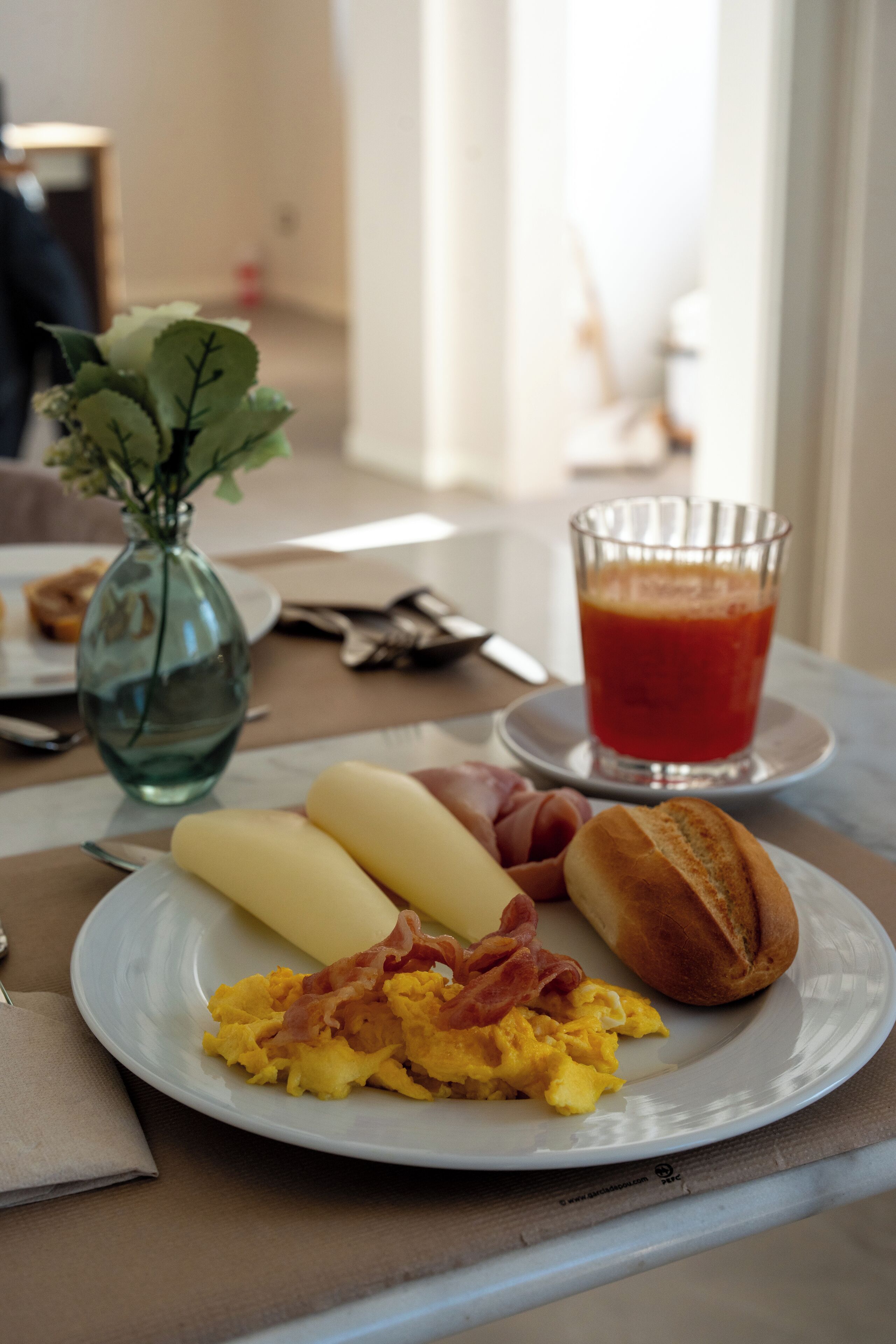 Ein Frühstücksteller mit Eiern, Speck und Toast auf einem Tisch, neben einer Blumenvase und einem Glas Orangensaft.
