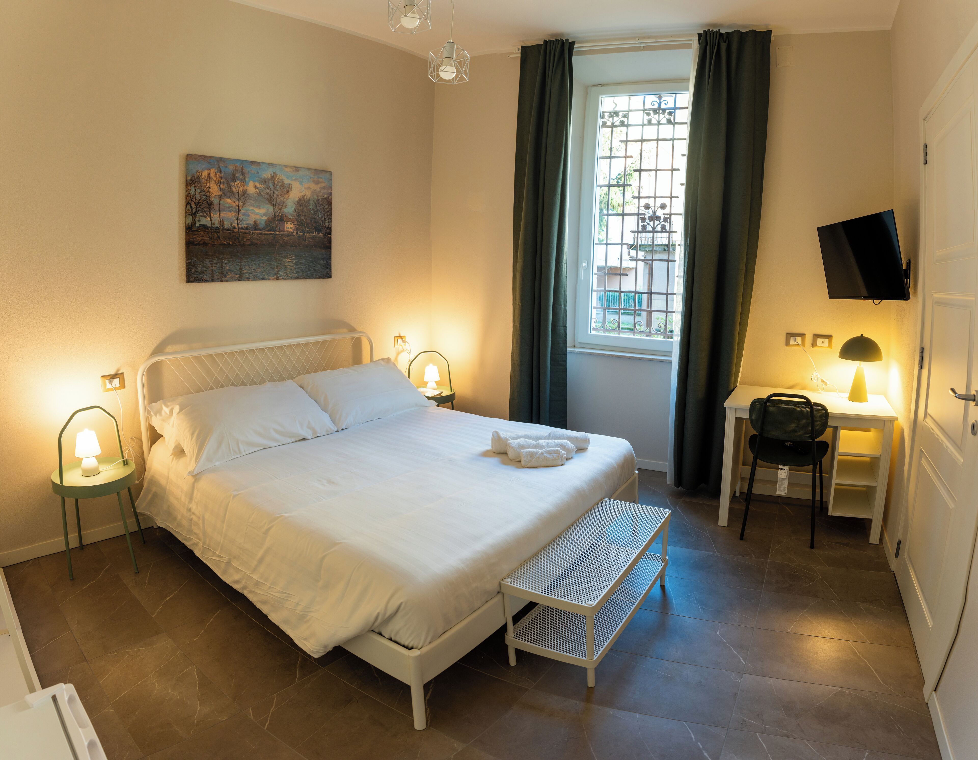 Ein ordentlich eingerichtetes Schlafzimmer mit Bett, Nachttischlampen, Schreibtisch mit Stuhl, Fernseher und einem Fenster mit Vorhängen.
