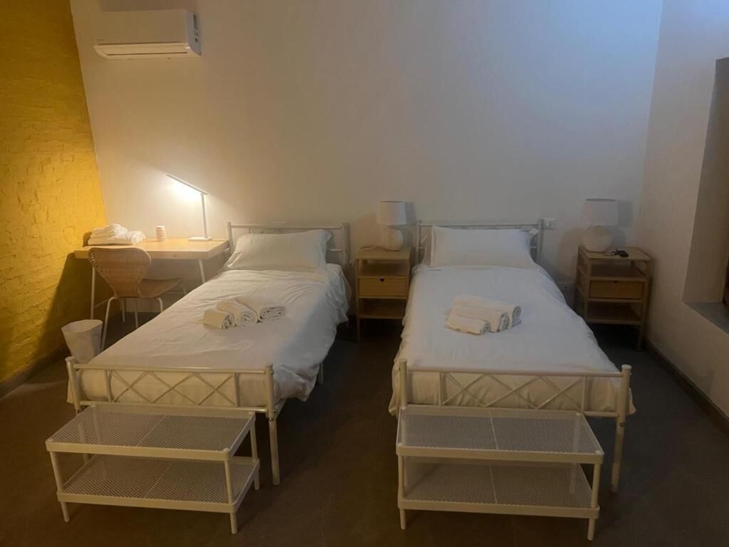 Habitación de hotel con dos camas individuales, cada una con mesita de noche y lámpara, un escritorio pequeño con silla y un cubo de basura.