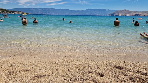 Una playa con agua turquesa cristalina, orilla de piedrecillas y montañas a lo lejos.