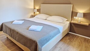 A neatly made bed with a grey quilt, two white pillows, and two folded white towels on the bed. There are two bedside tables with lamps, one on each side of the bed. - Charming Apartment Sunshine BS - with Balcony, 300 m from the Beach (Baska)