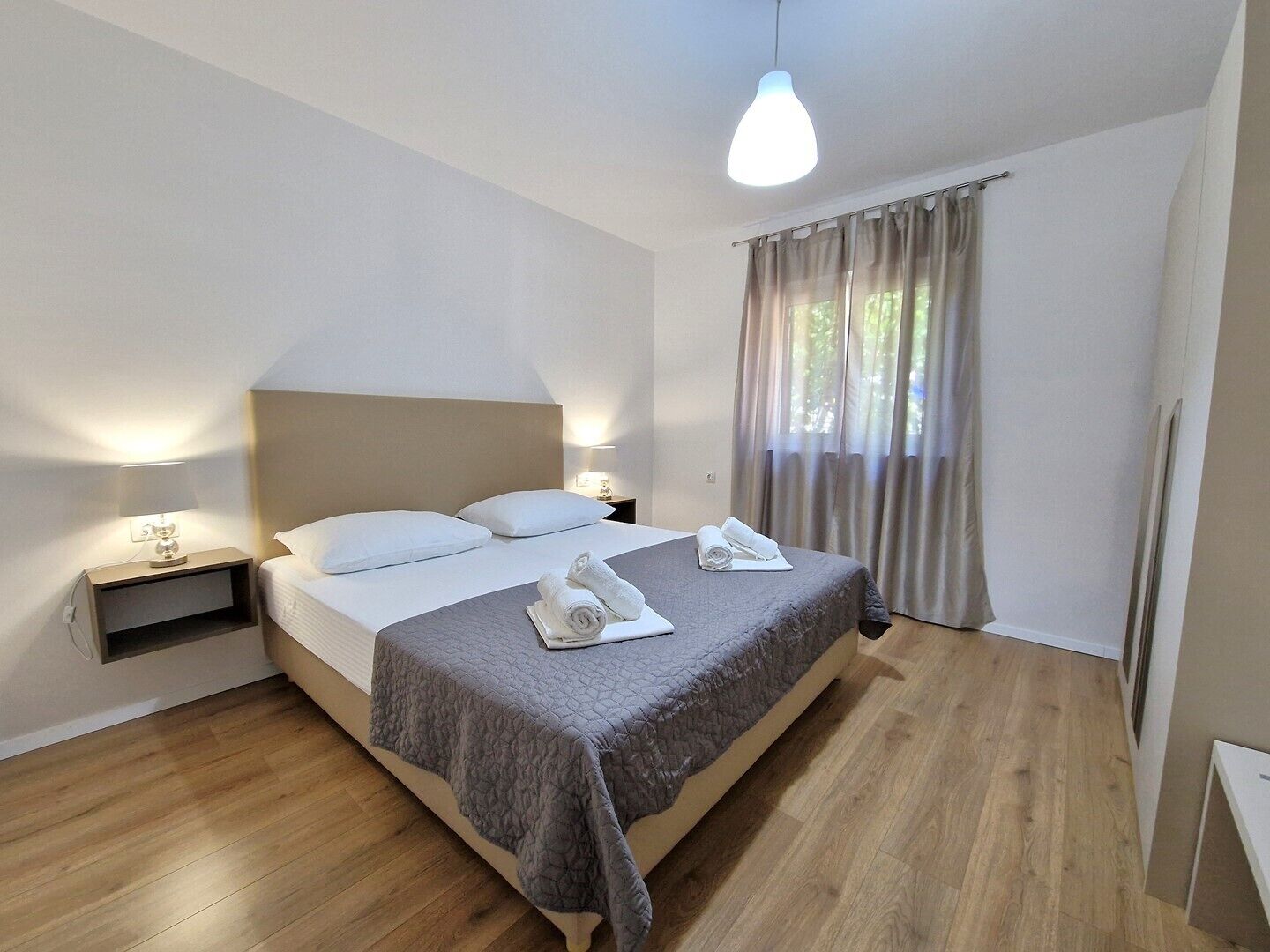 A modern hotel room with a large bed, two bedside tables, a bedside lamp, and a view of the outdoors through sheer curtains.