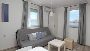 Een moderne woonkamer met een grijze bank, een klein wit tafeltje en een beige poef. Er zijn twee ramen met behang, een airconditioning en een wandlamp.
