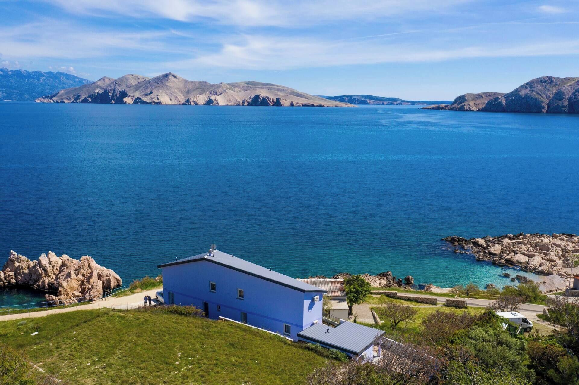 Une propriété située sur la côte, avec un bâtiment bleu, un rivage rocheux et des montagnes en arrière-plan.