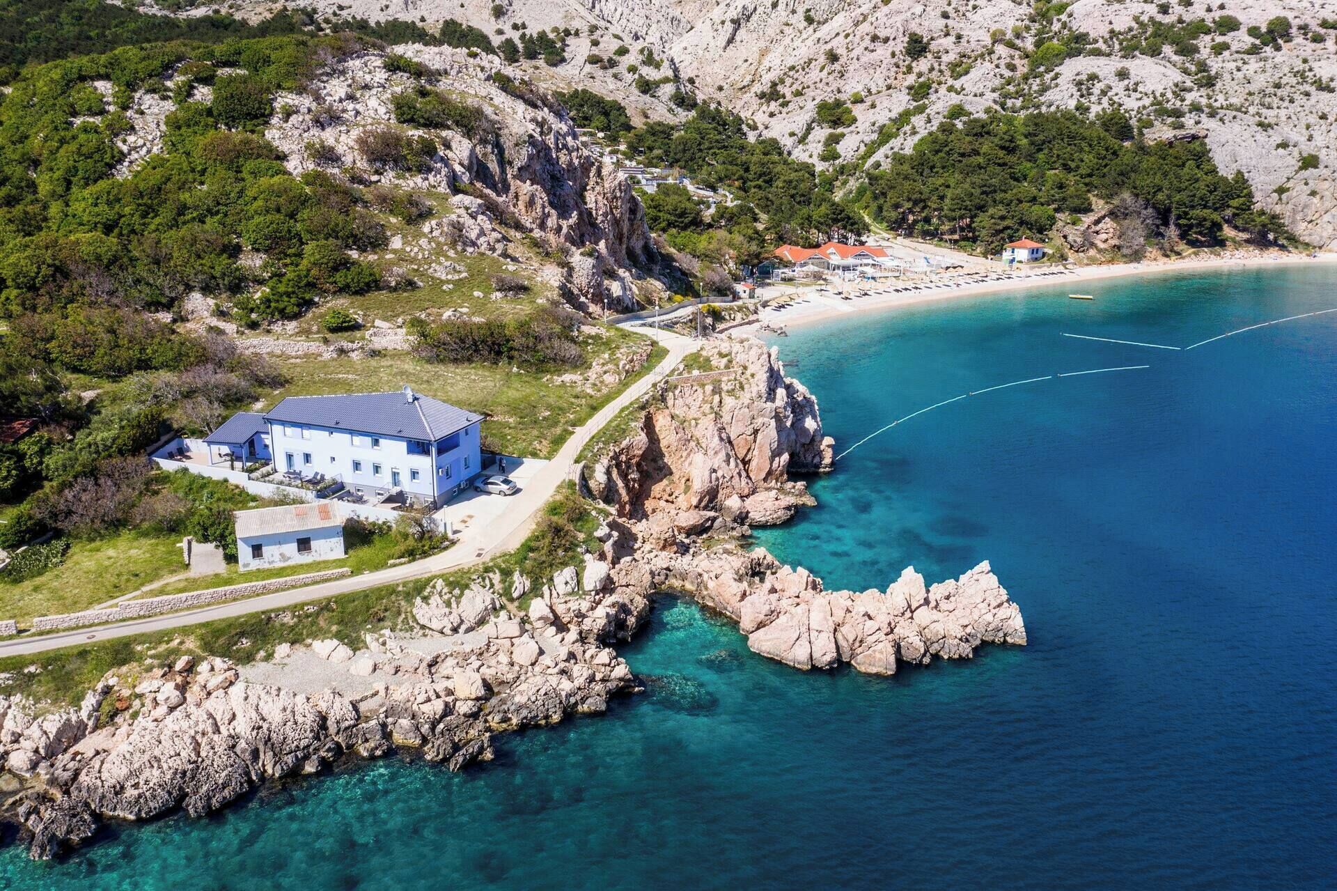 Vue aérienne d’une propriété située sur la côte, avec un grand bâtiment, une côte rocheuse et des eaux d’un bleu limpide.