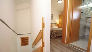 A hotel room with a wooden staircase, a bed with a headboard, a bathroom with a shower, and a small window.