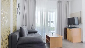 A modern living room with a grey sofa, a small wooden table, a TV on a stand, and a large window with sheer curtains.